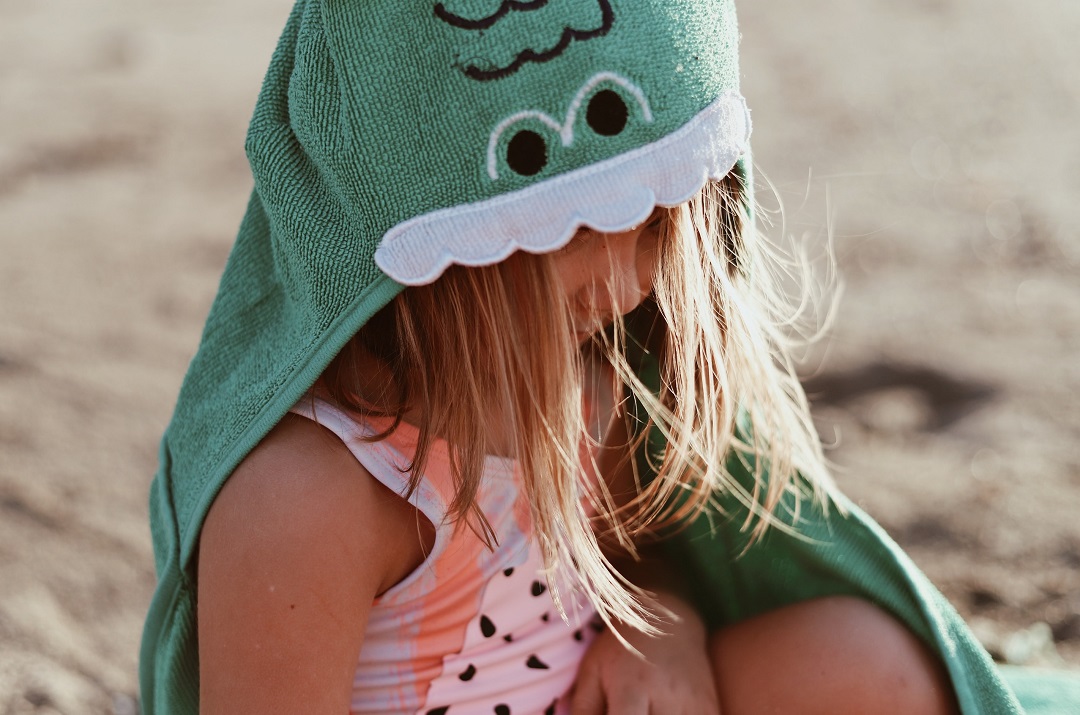 little girl sitting on beach with green towel over her head